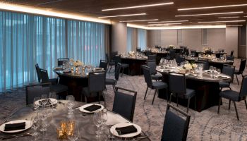 A banquet room with round tables set for an event. Each table has plates, glasses, napkins, floral centerpieces, and candles, with chairs neatly arranged.