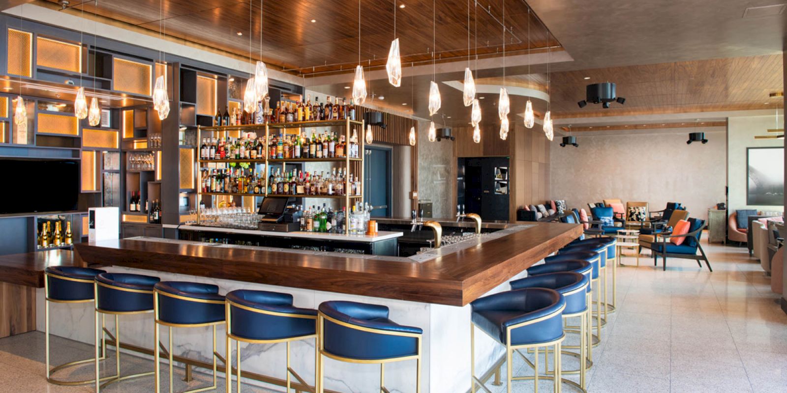 The image features a modern bar with a wooden counter, blue barstools, hanging lights, and a well-stocked shelf of liquor bottles in a contemporary setting.