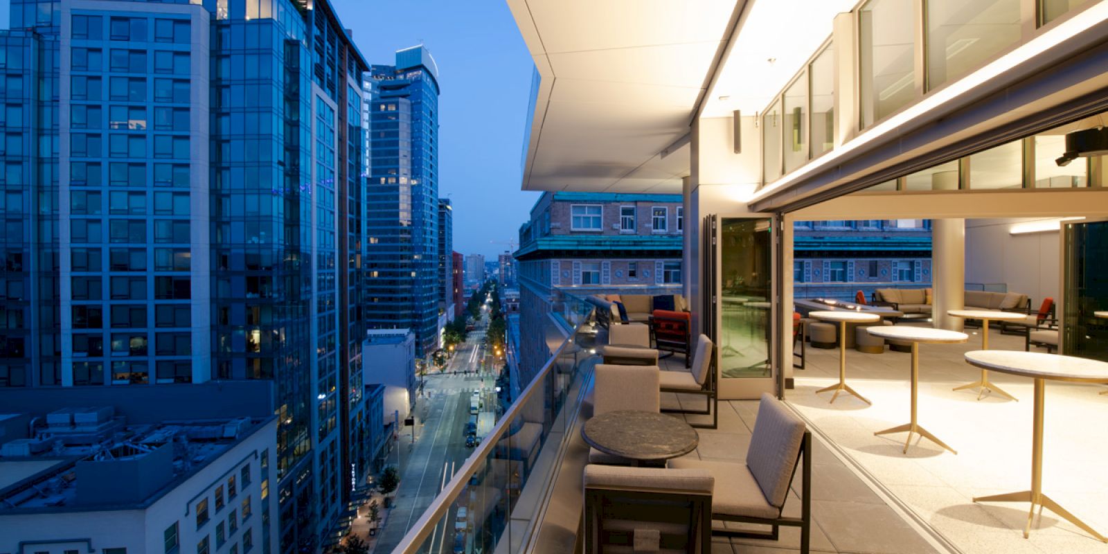 A modern cityscape with tall buildings at dusk, viewed from a balcony with tables and chairs, offering a luxurious and scenic view.
