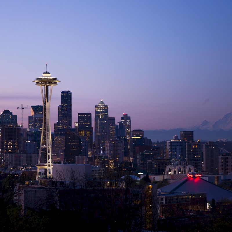 This image shows the skyline of Seattle at dusk, featuring the Space Needle prominently with Mount Rainier visible in the background.
