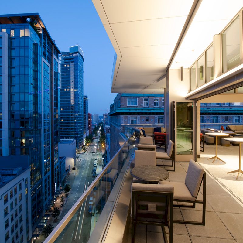 The image shows a modern cityscape at dusk, with a balcony view of tall buildings and a stylish outdoor seating area with tables and chairs.