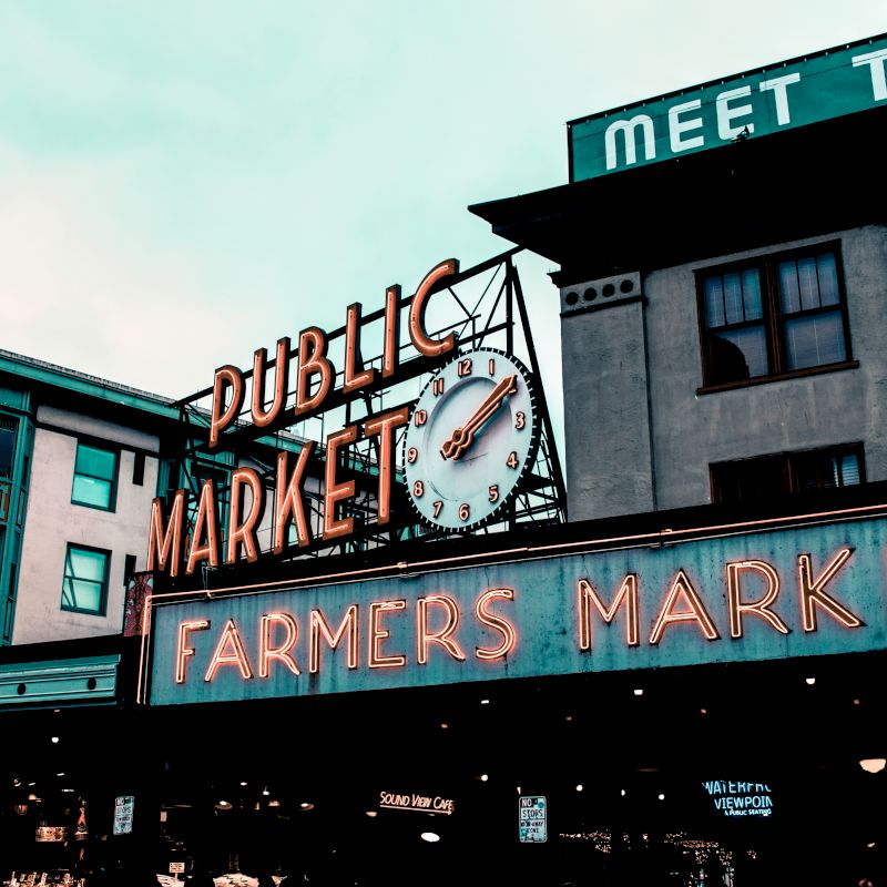 The image shows the iconic neon sign of a Public Market and Farmers Market, with the clock displaying the time, located in a busy urban setting.