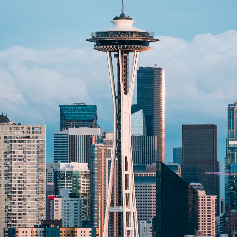 The image shows the Space Needle in Seattle, surrounded by various high-rise buildings and a partly cloudy sky in the background.