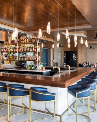 The image shows a modern bar with a wooden counter, blue cushioned barstools, pendant lights, and a fully stocked back bar, with seating areas in the background.