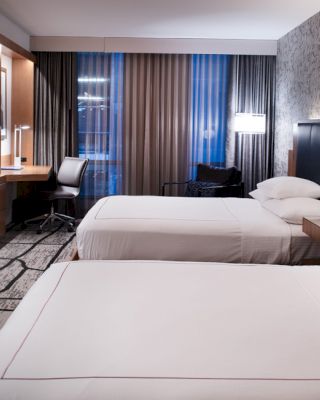 A modern hotel room with two beds, a wall-mounted TV, a desk, and dim lighting. The room features contemporary decor and a patterned accent wall.
