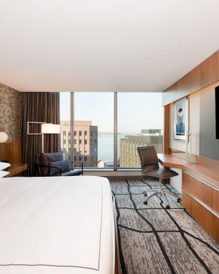 A modern hotel room with a king-size bed, a large TV, a desk, and a chair by the window offering a city view with buildings in the background.