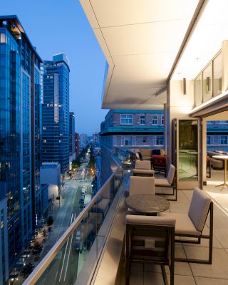 A modern cityscape viewed from a high-rise balcony at dusk, featuring outdoor seating and glass buildings illuminating the urban streets below.