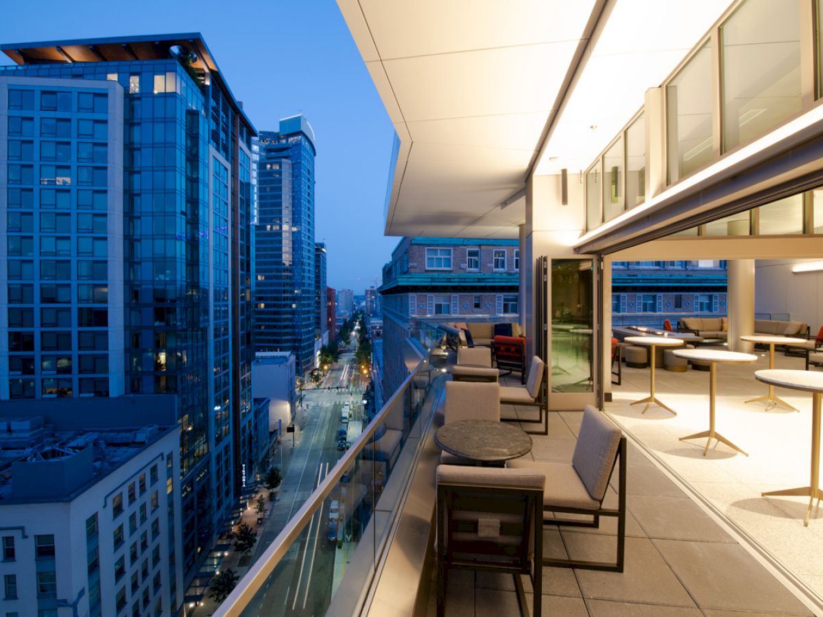 The image shows a modern rooftop patio with seating and tables, overlooking a cityscape with tall buildings and a street below during twilight.