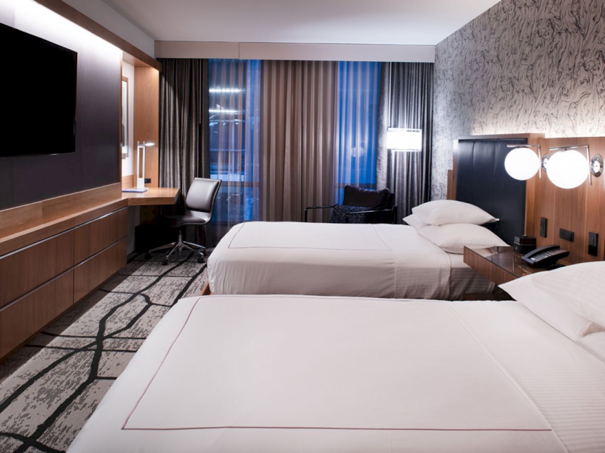 A modern hotel room with two double beds, mounted TV, desk, and stylish lighting on the wall. The decor includes patterned wallpaper and curtains.