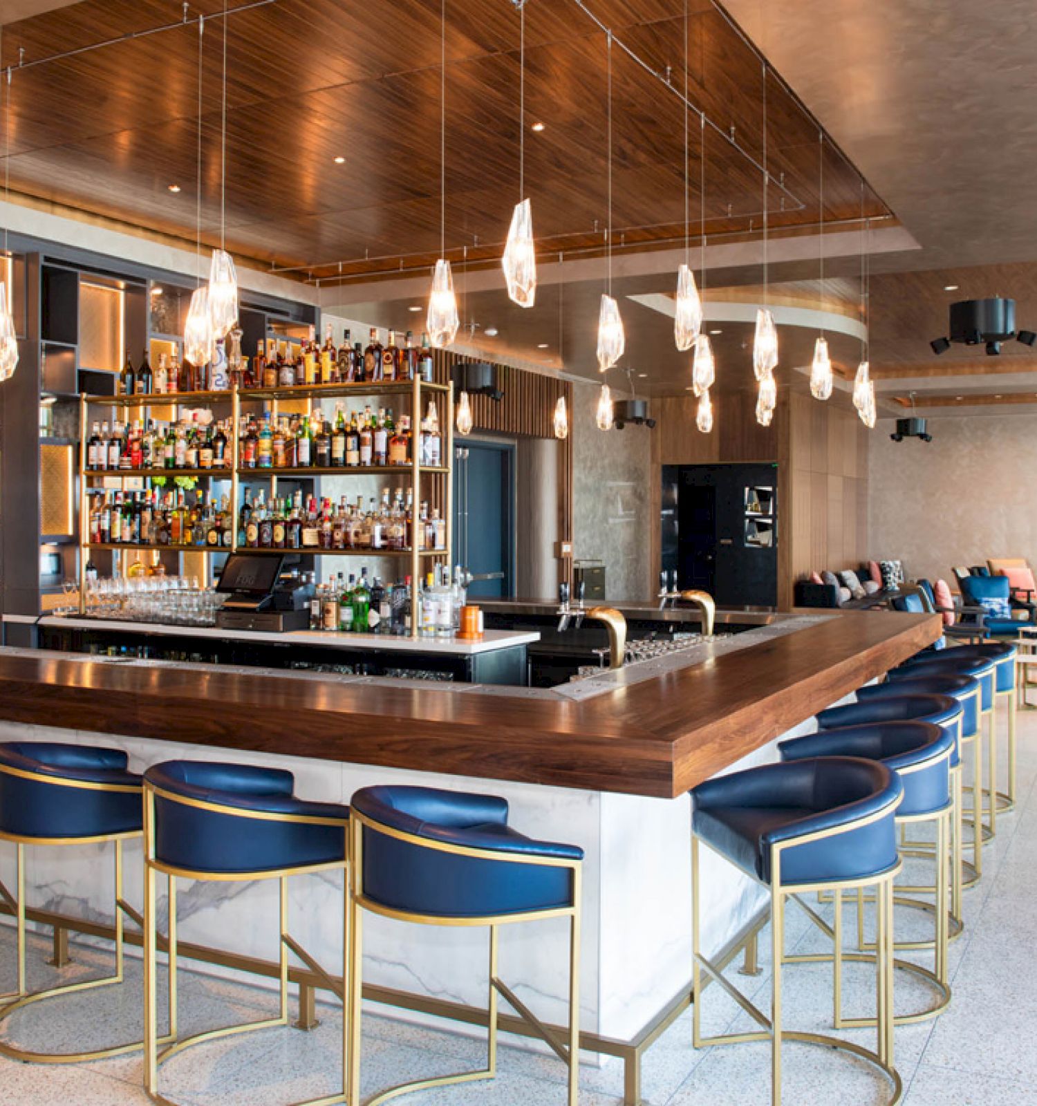 A modern bar with a well-stocked liquor shelf, blue bar stools, pendant lights, and a spacious seating area in the background.