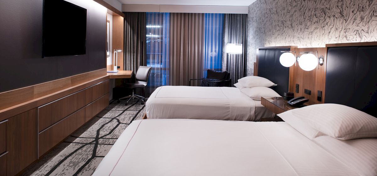 A modern hotel room with two neatly made beds, a flat-screen TV, a desk, and a chair; lights and curtains enhance the cozy atmosphere.
