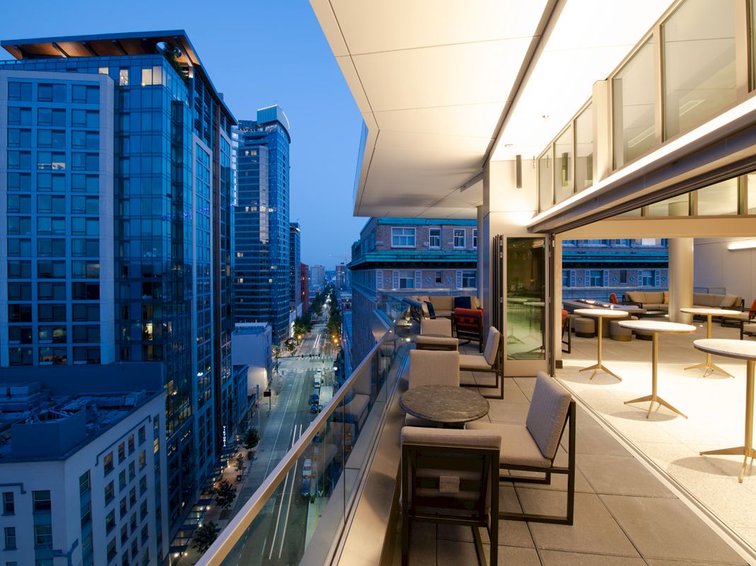 A modern urban balcony at dusk with outdoor seating overlooks a city street lined with tall buildings and lit windows, under a blue sky.