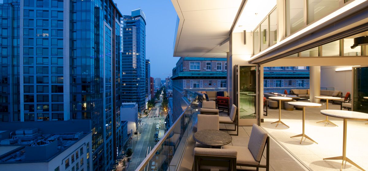 A modern urban balcony at dusk with outdoor seating overlooks a city street lined with tall buildings and lit windows, under a blue sky.