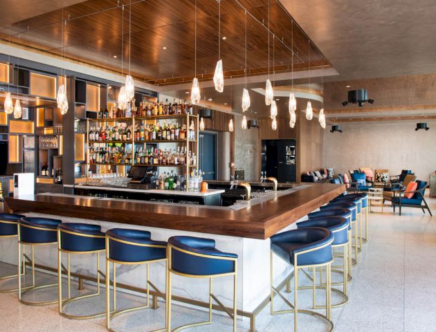 A modern bar with a large island counter, surrounded by blue and gold bar stools, under hanging lights. Shelves are stocked with various bottles.