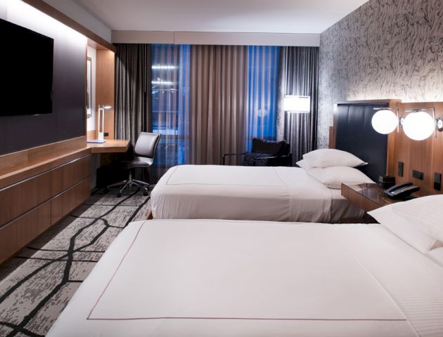 The image shows a modern hotel room with two neatly made beds, a wall-mounted TV, a desk with a chair, and stylish lighting.