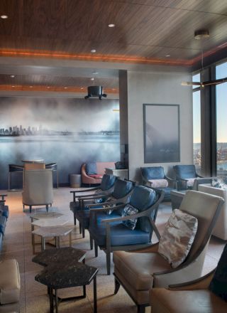 A modern lounge with plush seating, large windows offering a scenic water view, and a wall mural depicting a city skyline in the distance.