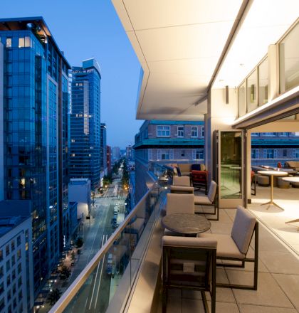 This image shows a modern, city view from a high-rise balcony at dusk with outdoor seating, tables, and illuminated buildings in the background.