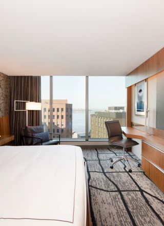 The image shows a modern hotel room with a king-size bed, large TV, desk, chair, and a view of buildings through large windows ending the sentence.