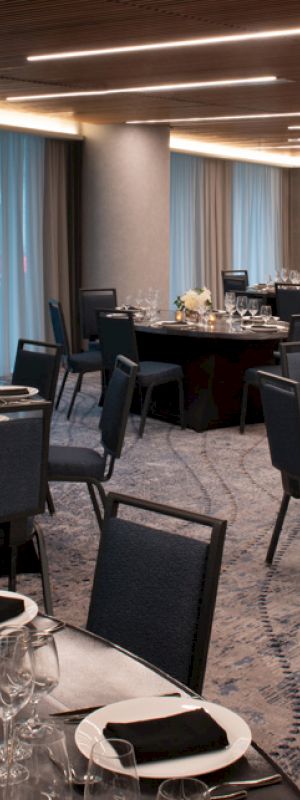 A modern, well-lit banquet hall with round tables set for an event, adorned with black napkins, white flowers, and candles, and featuring blue drapes.
