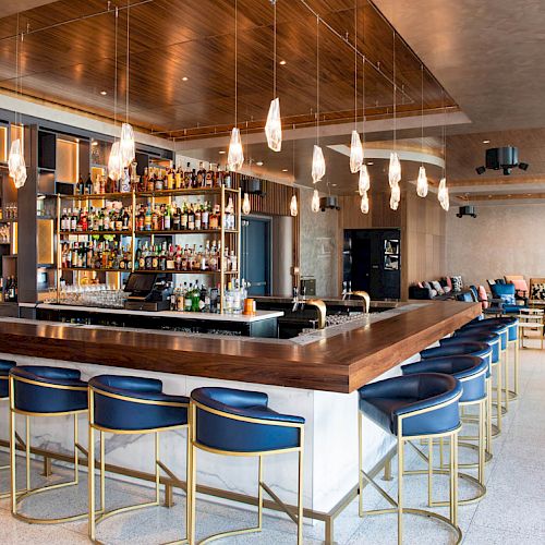 This image shows a modern bar with blue barstools, a well-stocked bar, pendant lights, and a spacious seating area with tables and chairs.