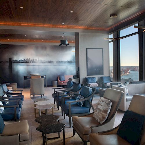 A modern lounge area with comfortable seating, large windows with a scenic view, and stylish decor including wall art and ceiling lighting.