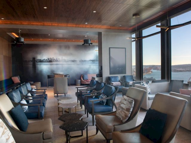 A lounge with comfortable seating, tables, and large windows showing a city view.