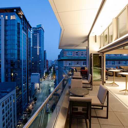 This image shows a modern balcony with seating, overlooking a bustling cityscape of tall buildings and illuminated streets at dusk.