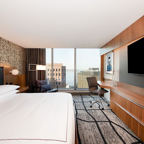 A modern hotel room features a large bed, desk with chair, wall-mounted TV, and a window offering a city view.