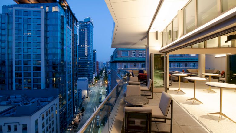 A balcony overlooking a city street with tall buildings, featuring outdoor seating and a modern indoor area.