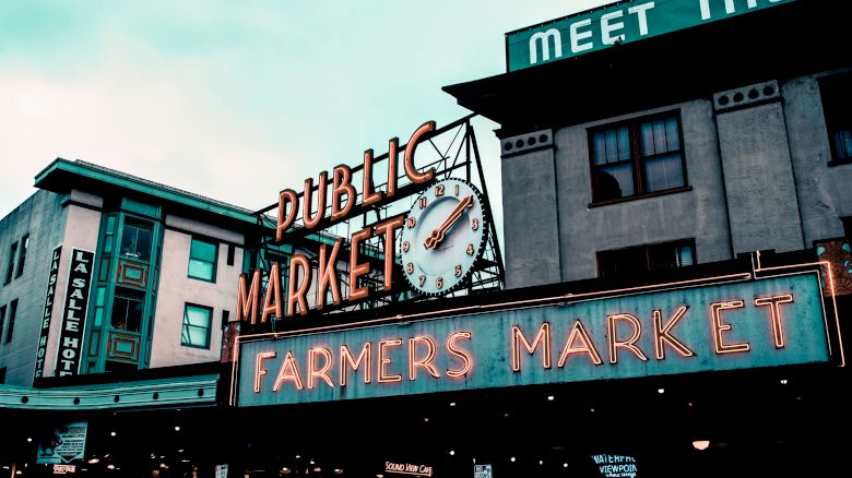 The image shows neon signs for a "Public Market" and "Farmers Market" on buildings, along with a clock and additional signs above the area.