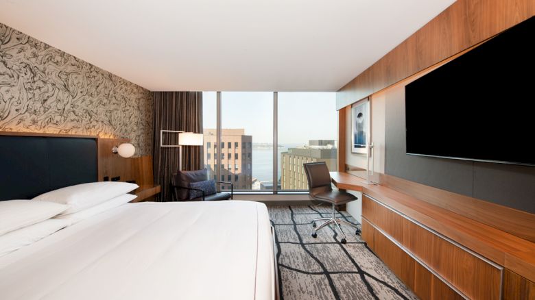 A modern hotel room with a large bed, wall-mounted TV, desk, chair, and a view of buildings and water through a large window.