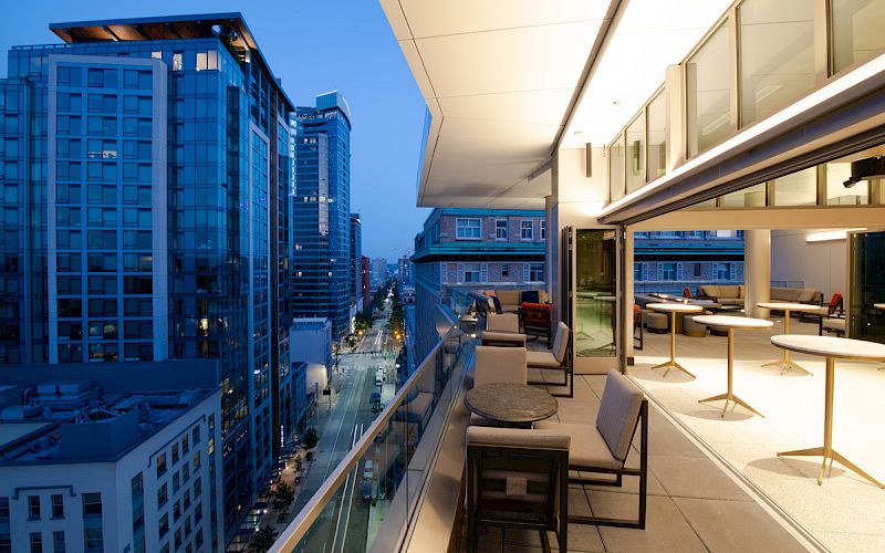 A rooftop patio with chairs, tables, and a glass railing overlooking a cityscape with tall buildings at dusk.
