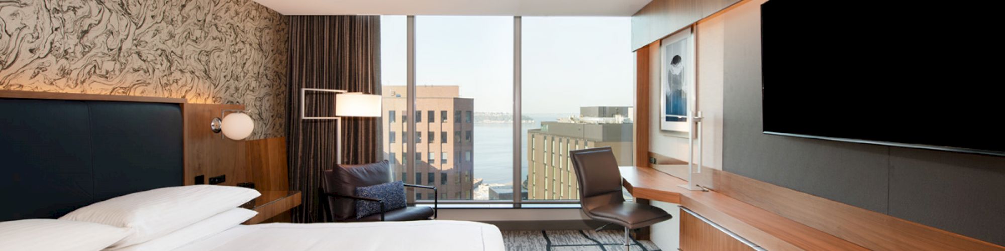 A modern hotel room with a large bed, a flat-screen TV, a desk with a chair, and a window with a city view.