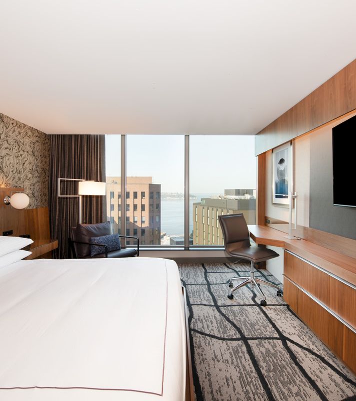 A hotel room with a bed, desk, chair, and TV, and a city view through the window.