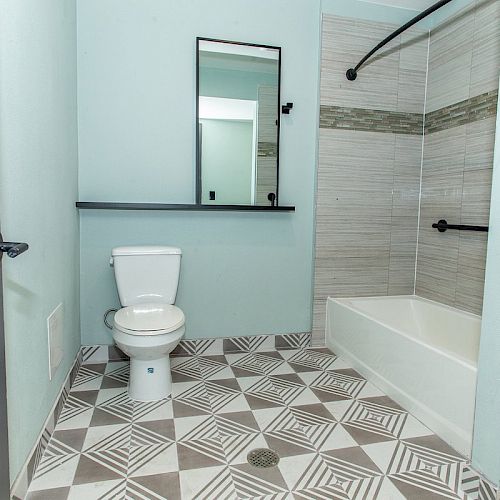 A bathroom featuring a toilet, a mirror, patterned floor tiles, and a bathtub-shower combo with tile walls and a curved shower curtain rod.