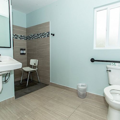 A clean, accessible bathroom with a sink, mirror, toilet, shower area with a chair, a grab bar, and a window, all set in a light-colored room.