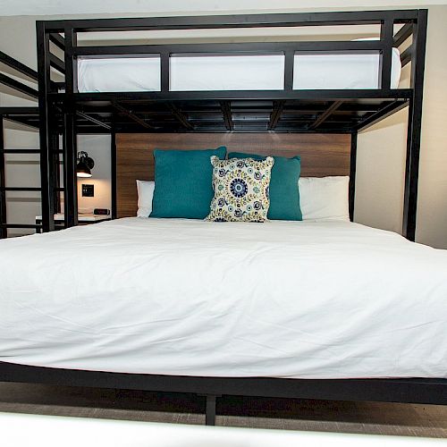 A bedroom with a large bed featuring white bedding and three decorative pillows under a bunk bed, with a window and an air conditioning unit.