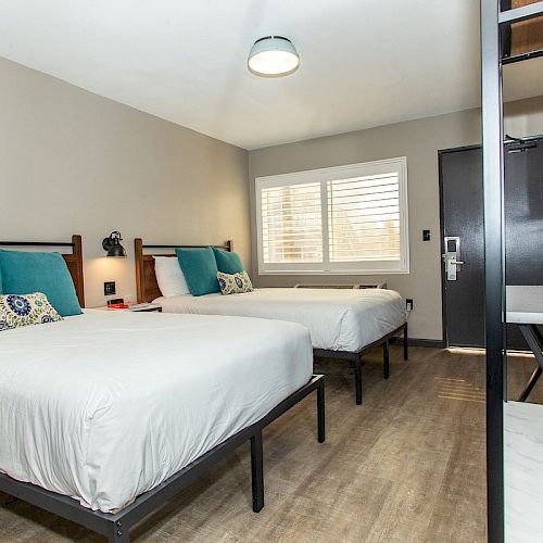 The image shows a clean, modern hotel room with two neatly made beds, teal cushions, a window with blinds, and minimalistic furniture.