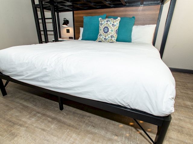 A close-up of a bed with white bedding and teal pillows in a room with a wooden headboard and metal frame.