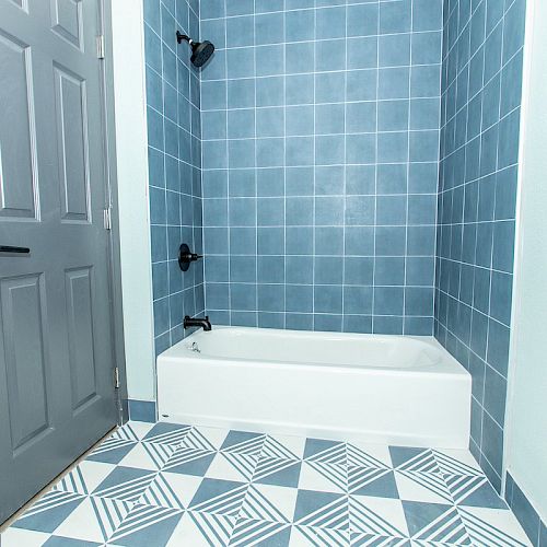 The image shows a bathroom with a blue tiled wall, a white bathtub, and a patterned blue and white floor. A gray door is seen to the left.