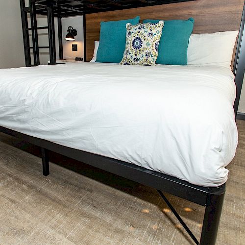 A neatly made bed with white linens, three decorative pillows against a dark headboard, and a wooden floor in the background.