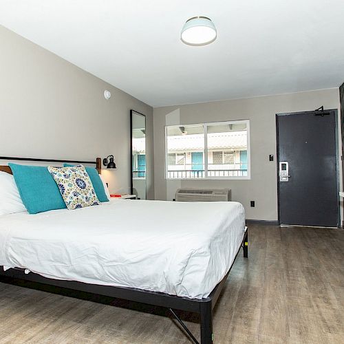 A modern hotel room features a bed with colorful pillows, two side tables, a mirror, a desk, a chair, and a window by the entrance door.