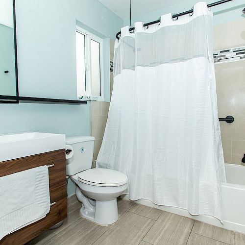 The image shows a modern bathroom with a sink, toilet, and bathtub with a white curtain. The room has light-colored walls and wooden flooring.