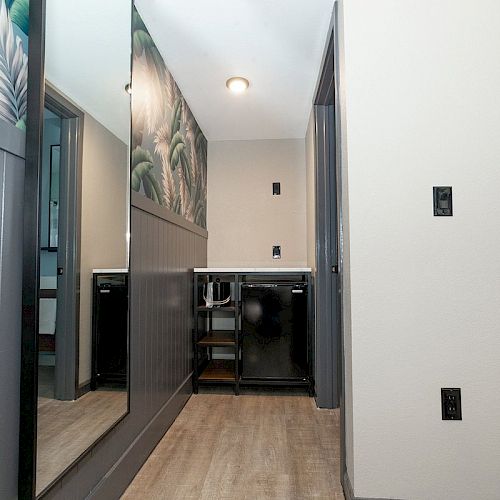 The image shows a hallway with a large mirror on the left wall, tropical wallpaper, a counter at the end, and a light fixture on the ceiling.