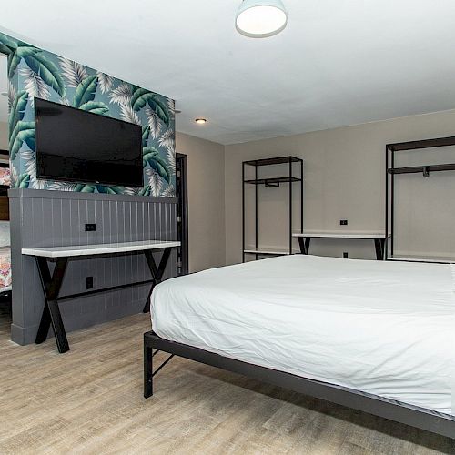 A modern room features a large bed, a TV mounted on the wall with tropical wallpaper, bunk beds, and minimalist black frame furniture.