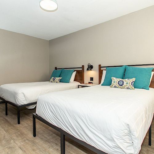 The image shows a hotel room with two neatly-made beds, each having white sheets and teal cushions, minimalist decor, and wood flooring.