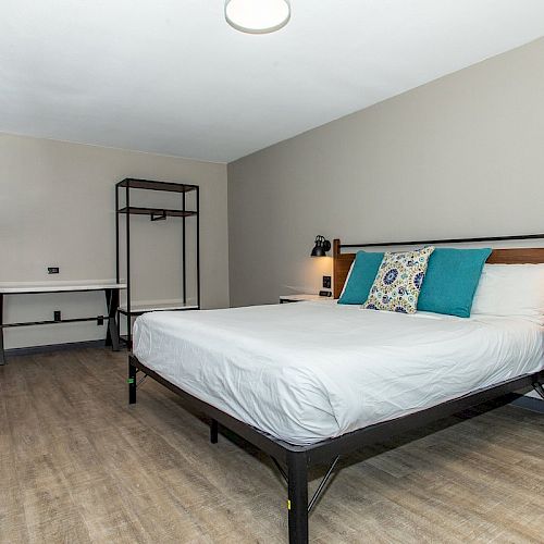 The image shows a minimalist bedroom with a large bed, a desk with chairs, shelves, and a light wooden floor, giving a clean and modern look.