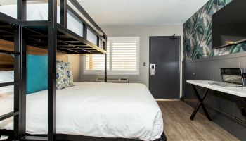 The image shows a modern hotel room with a bunk bed over a double bed, a TV on the wall, a desk with a laptop, and a window with white shutters.