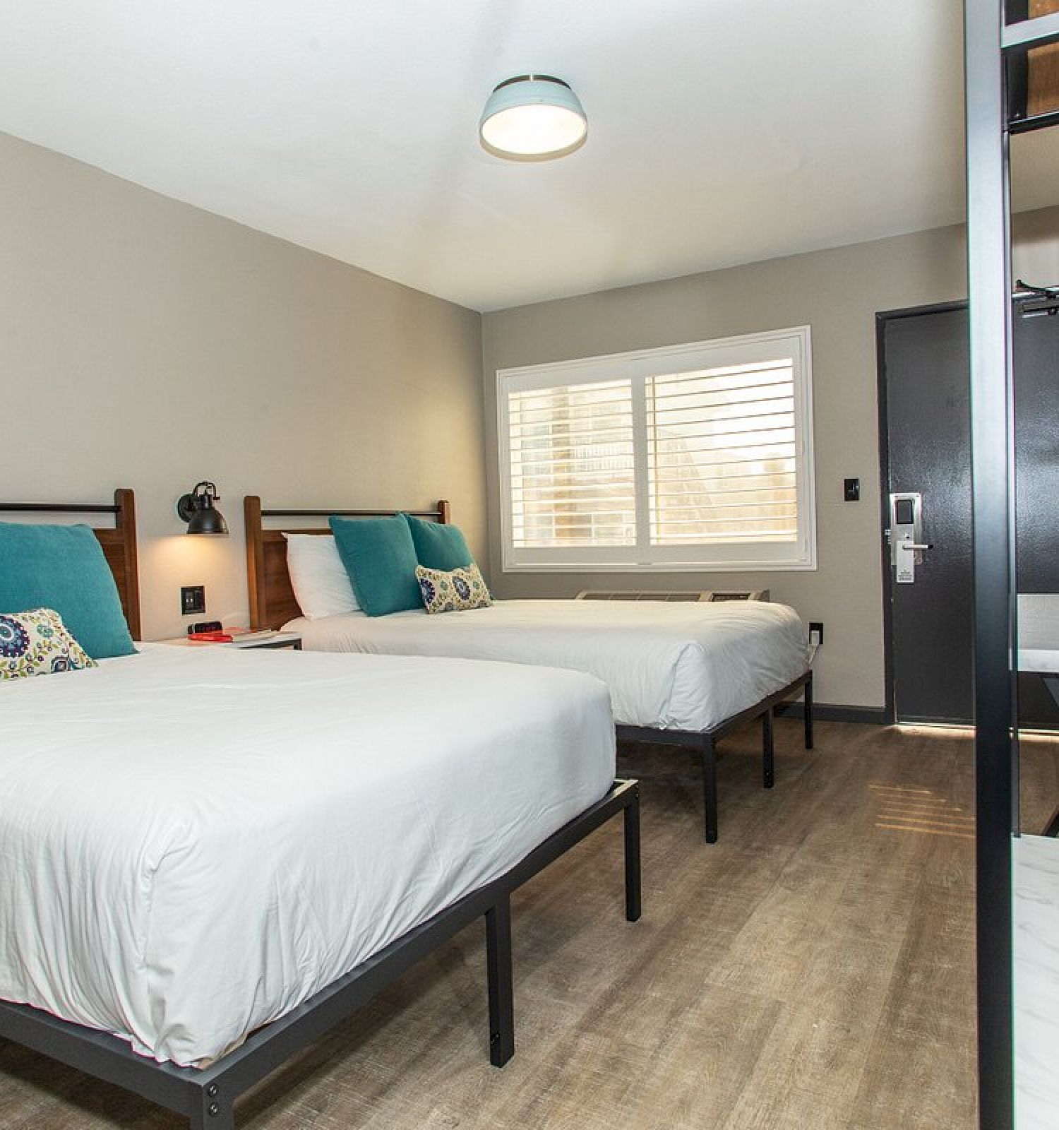 This image shows a modern hotel room with two neatly made beds, blue pillows, a large window, and minimalist decor, featuring wooden and black accents.