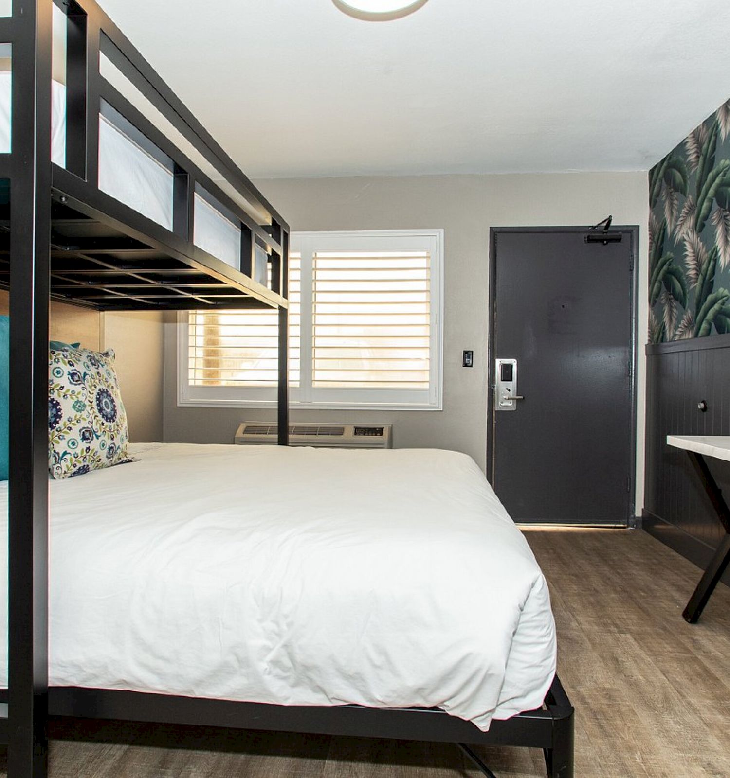 A modern hotel room with bunk beds, a large bed, desk with laptop, TV, palm-leaf wallpaper, and a door with keycard access in the background.